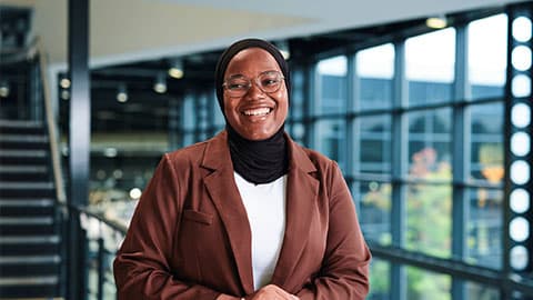 Mubeenat is standing and smiling and in focus. She is wearing a brown blazer and white t-shirt. In the background are big windows which are blurred.